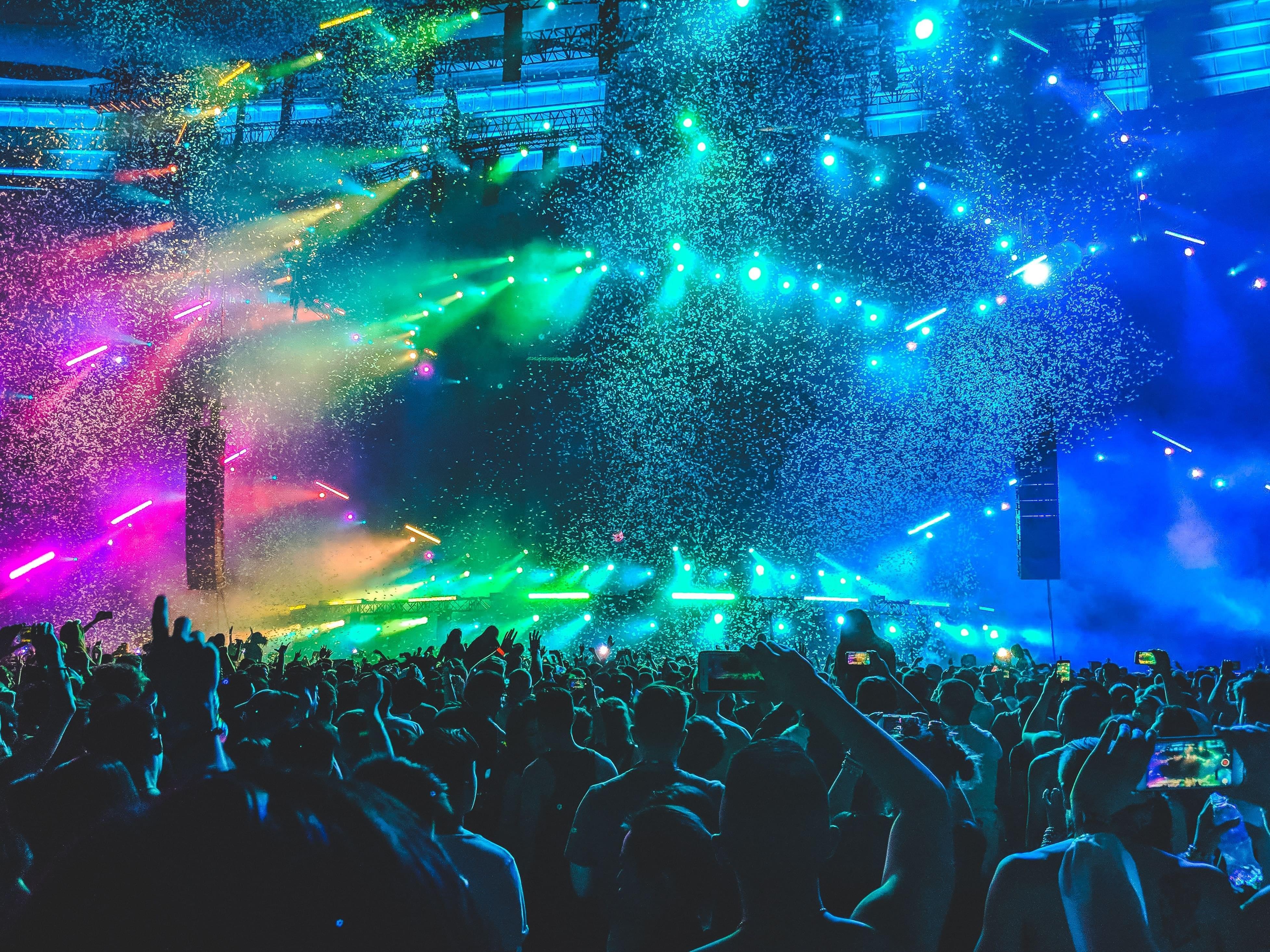 A large crowd facing a stage at a music festival with rainbow spectrum spotlights and confetti