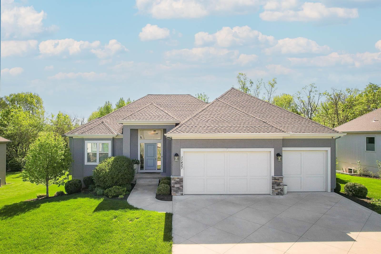 Front exterior of home for sale at 11427 S Redbud Lane Olathe KS — gray stucco reverse 1.5-story with 3-car garage, stone accents, and manicured landscaping.