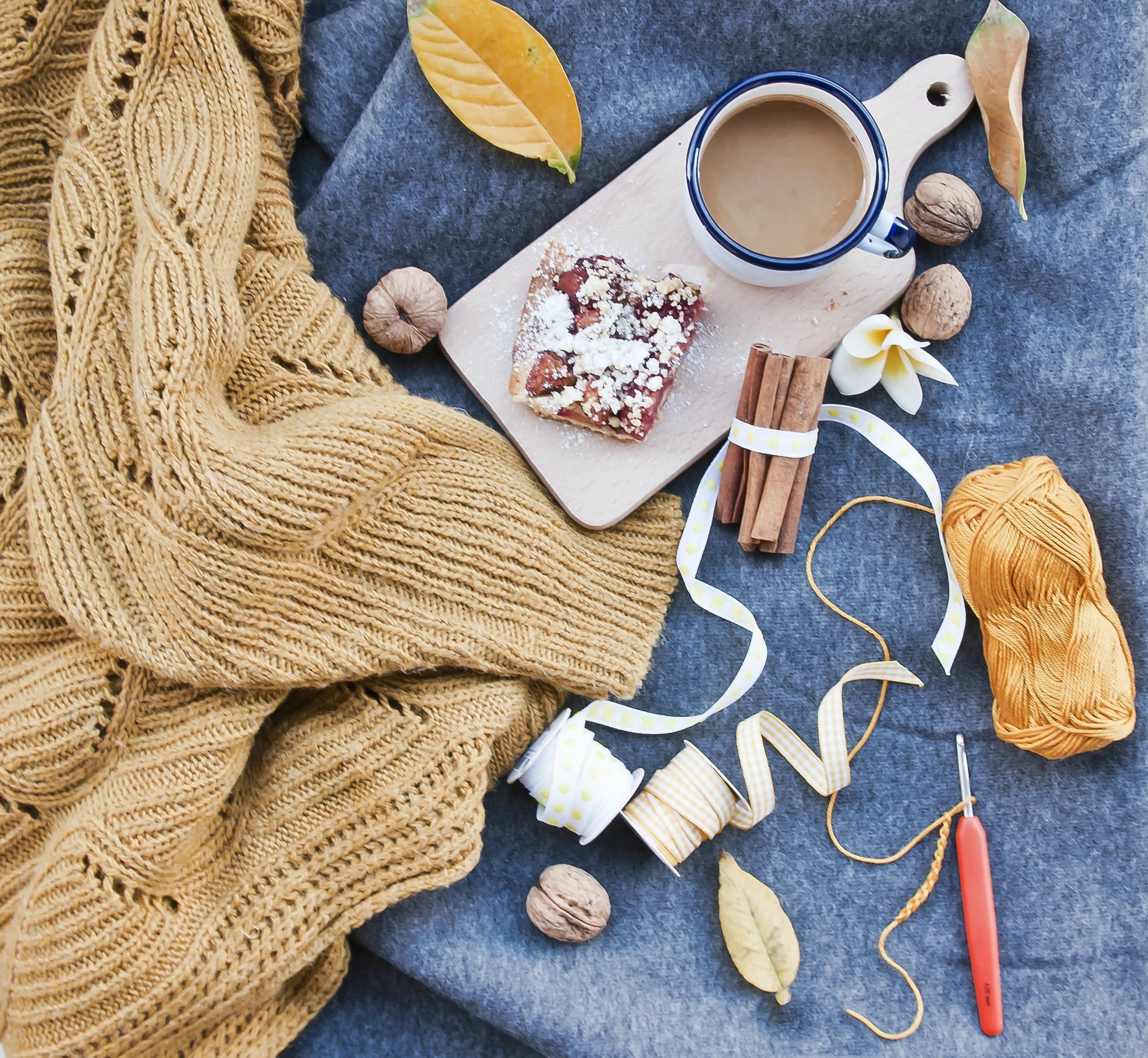 A blue felt surface with a beige sweater, gold yan, white and cream ribbon, crochet hook, and fall treats