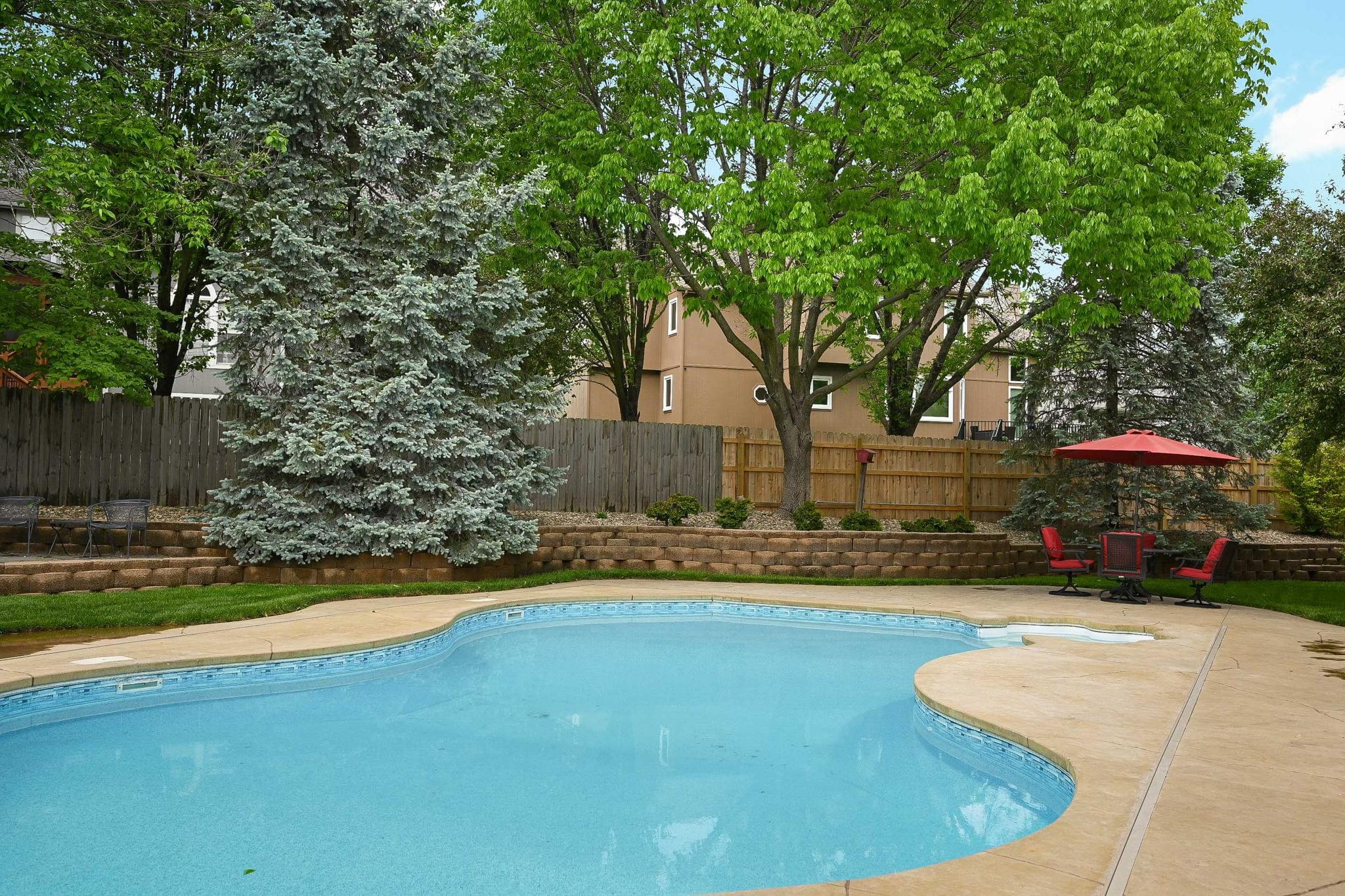 In-ground pool at 14730 S Hallet St in Olathe, KS, with mature trees, patio seating, retaining wall, and fenced backyard.