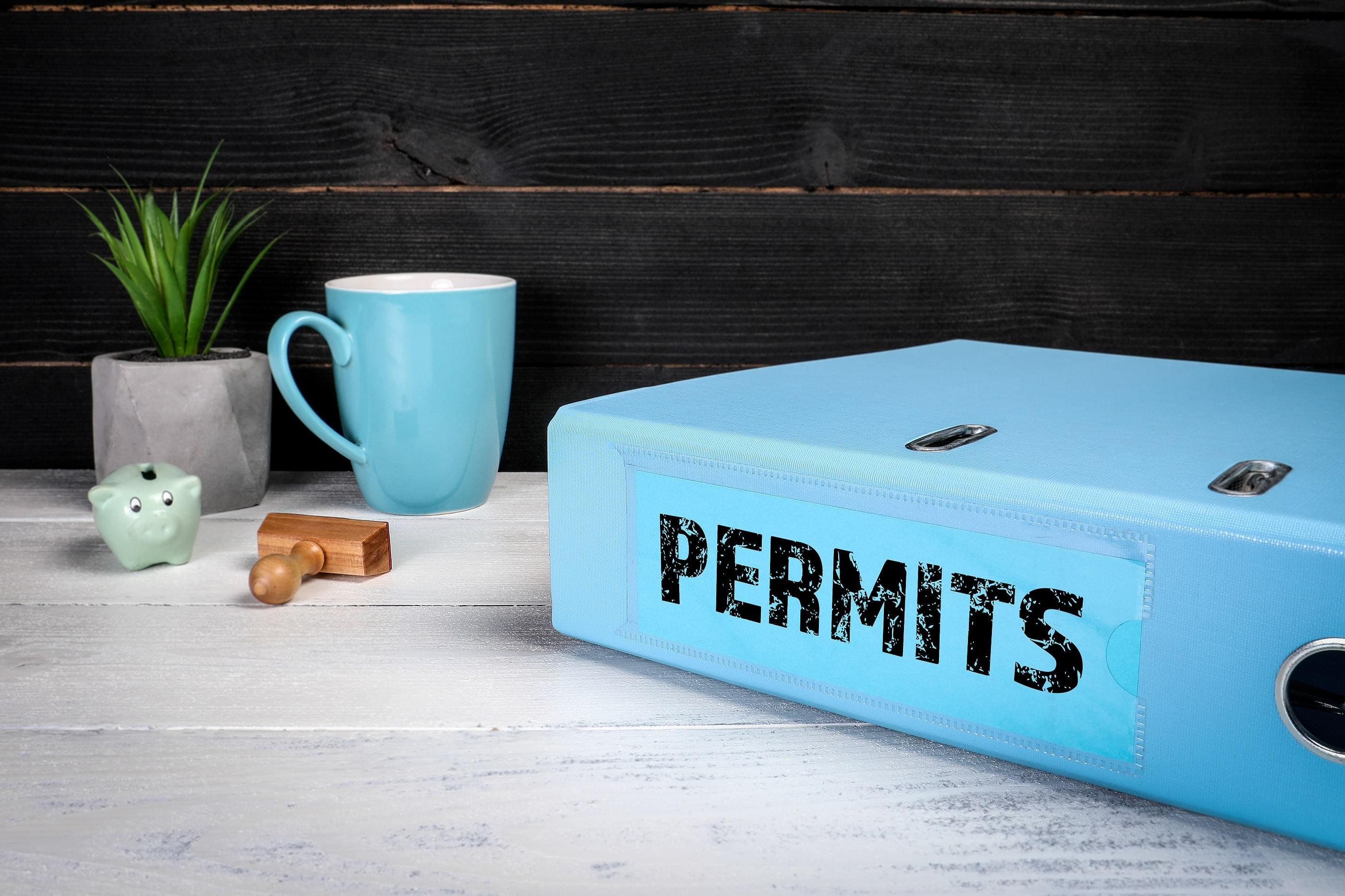 A blue binder labeled "Permits" on a table with a plant, a blue coffee cup, a piggy bank, and a stamp.