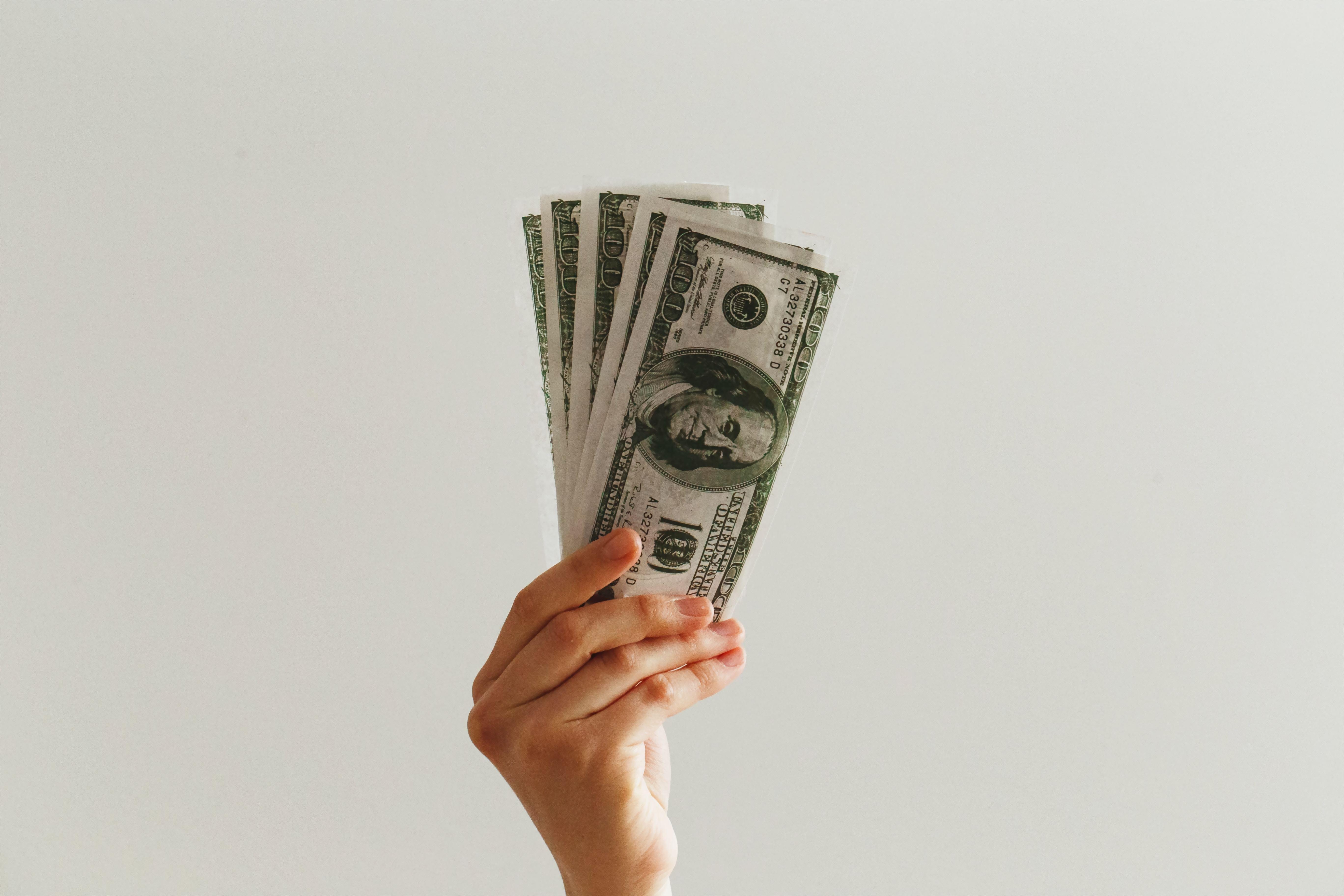 A hand holding several 100-dollar bills in front of an off-white background