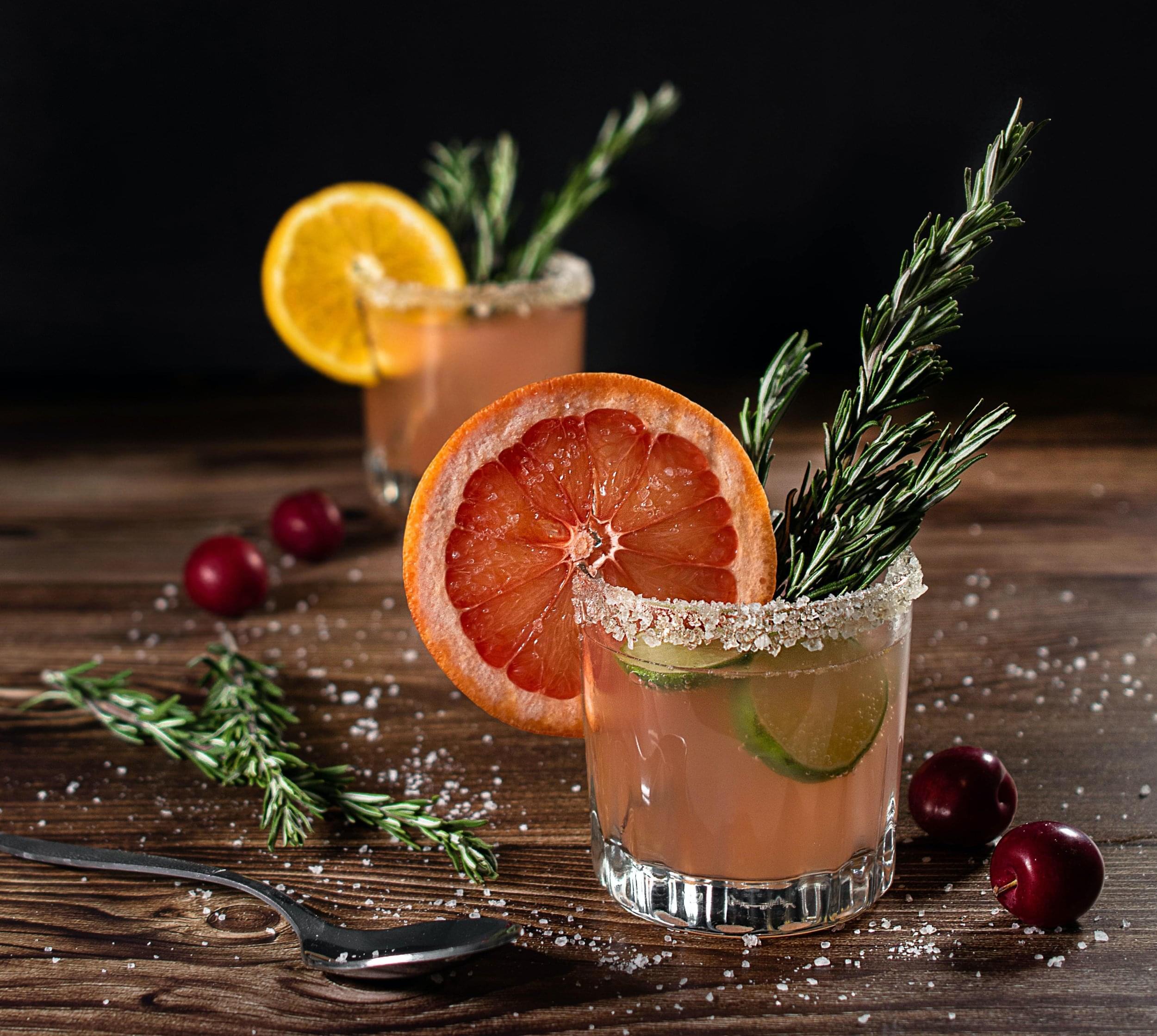 Two fancy cocktails with sugar on the rim, sage garnish, berries, and large citrus slices on a wooden table with a black background