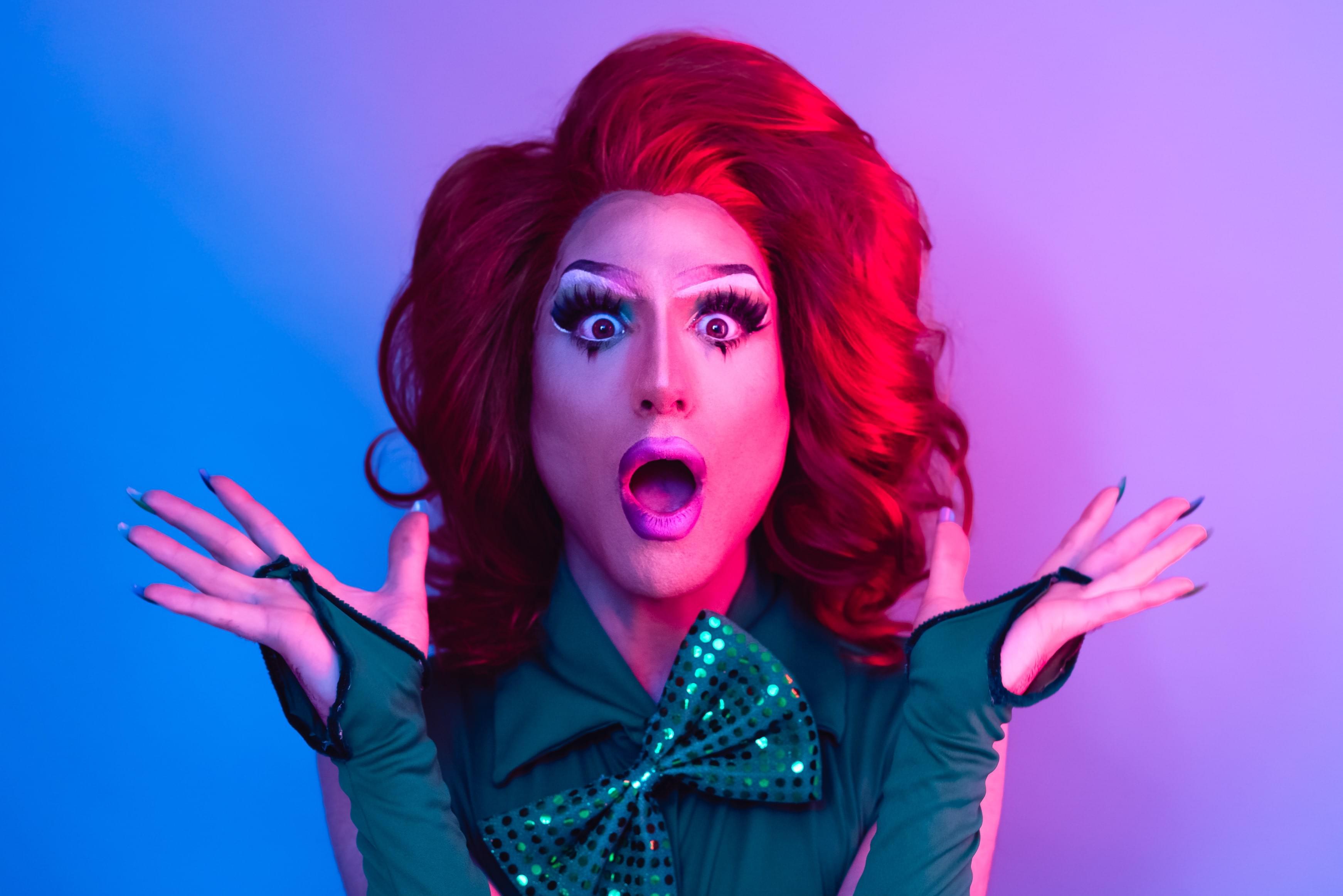 A red-haired drag queen looking surprised in front of a blue and purple background.