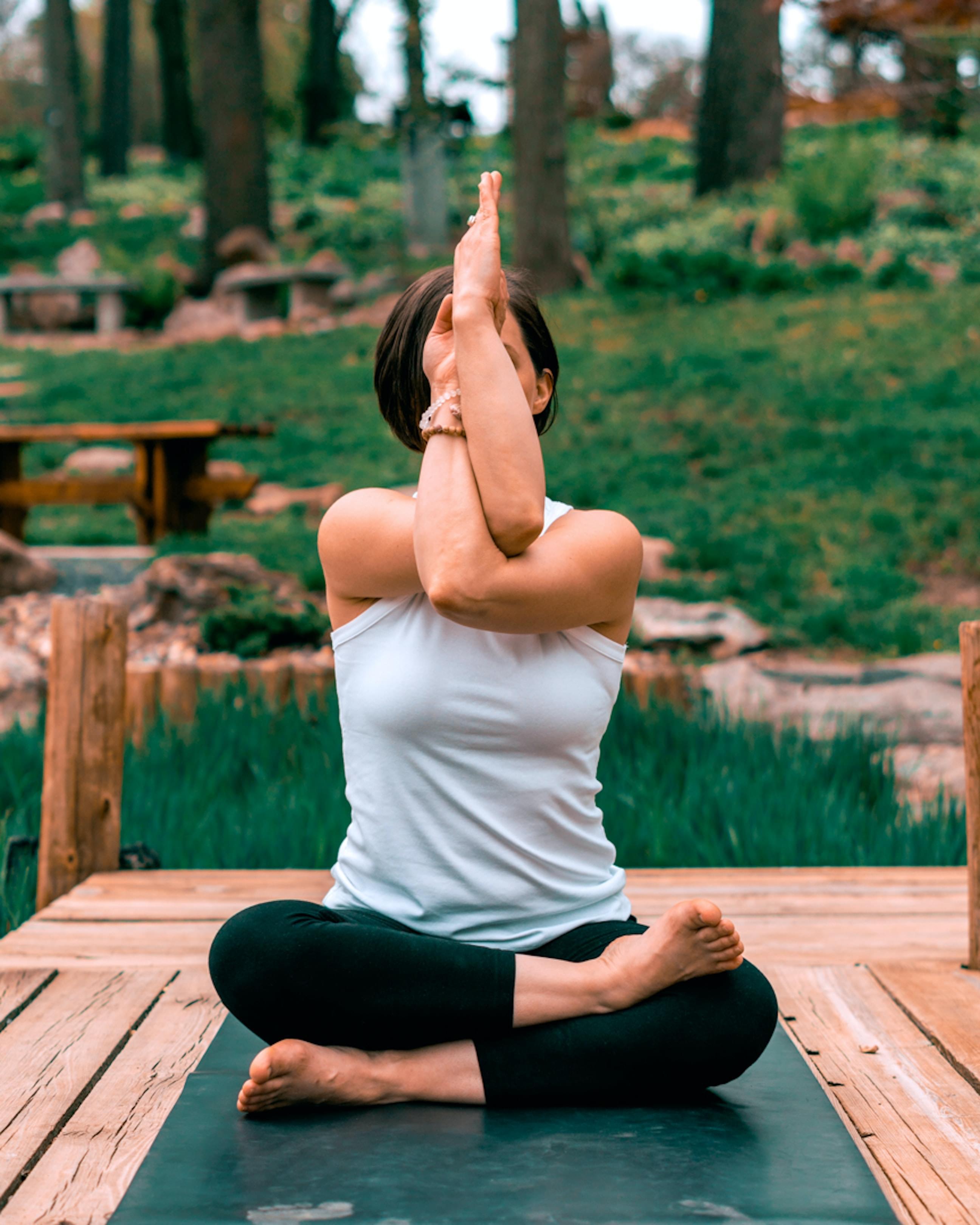 A dark-haired woman doing a seated yoga pose with arms twisted in front of her face on a wooden deck in front of a green space