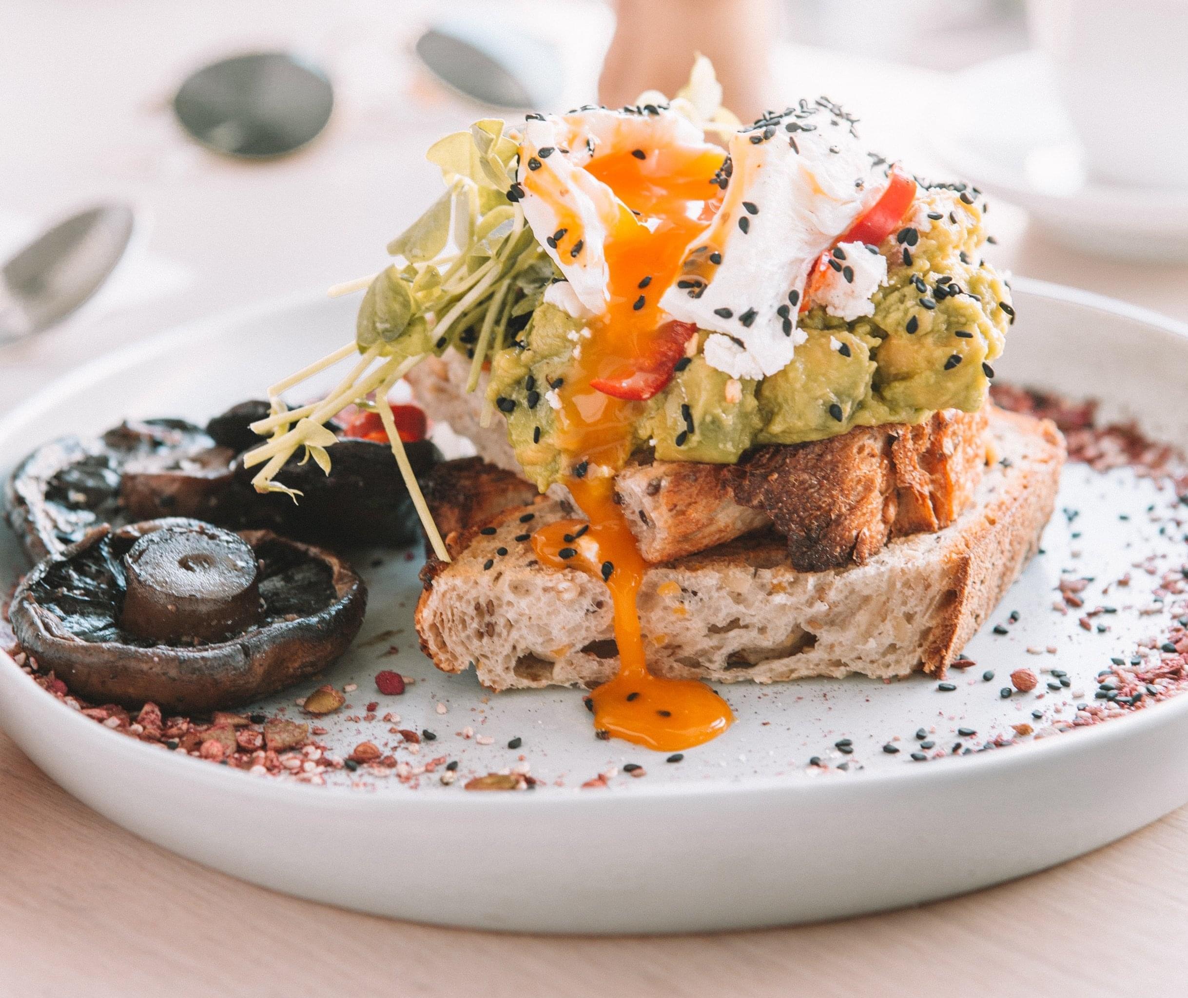 A brunch plate featuring egg, avocado, chia seats, and sprouts on toast with mushrooms on the side