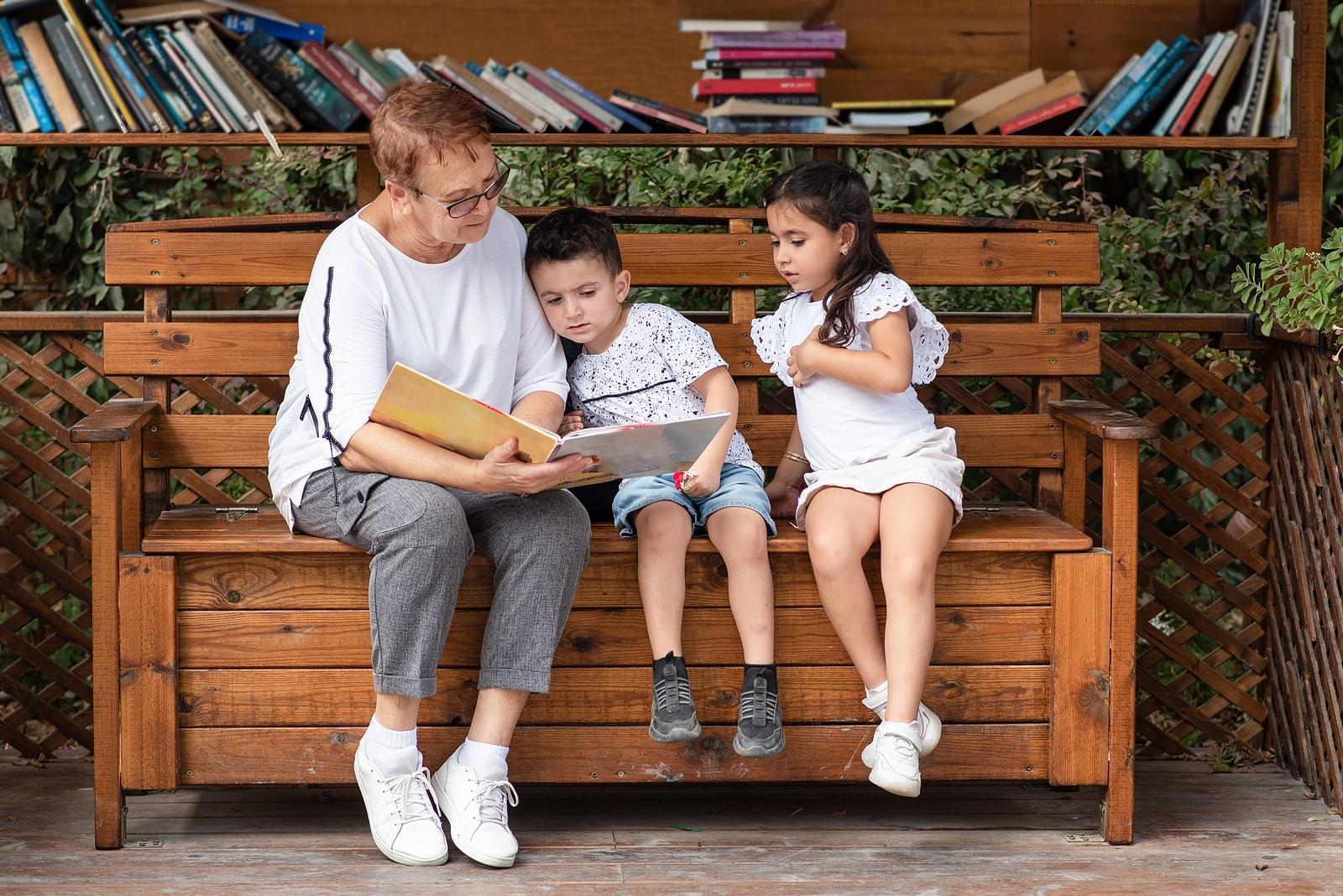A grandmother reading a book with two children on a wooden bench at a library in front of a book shelf