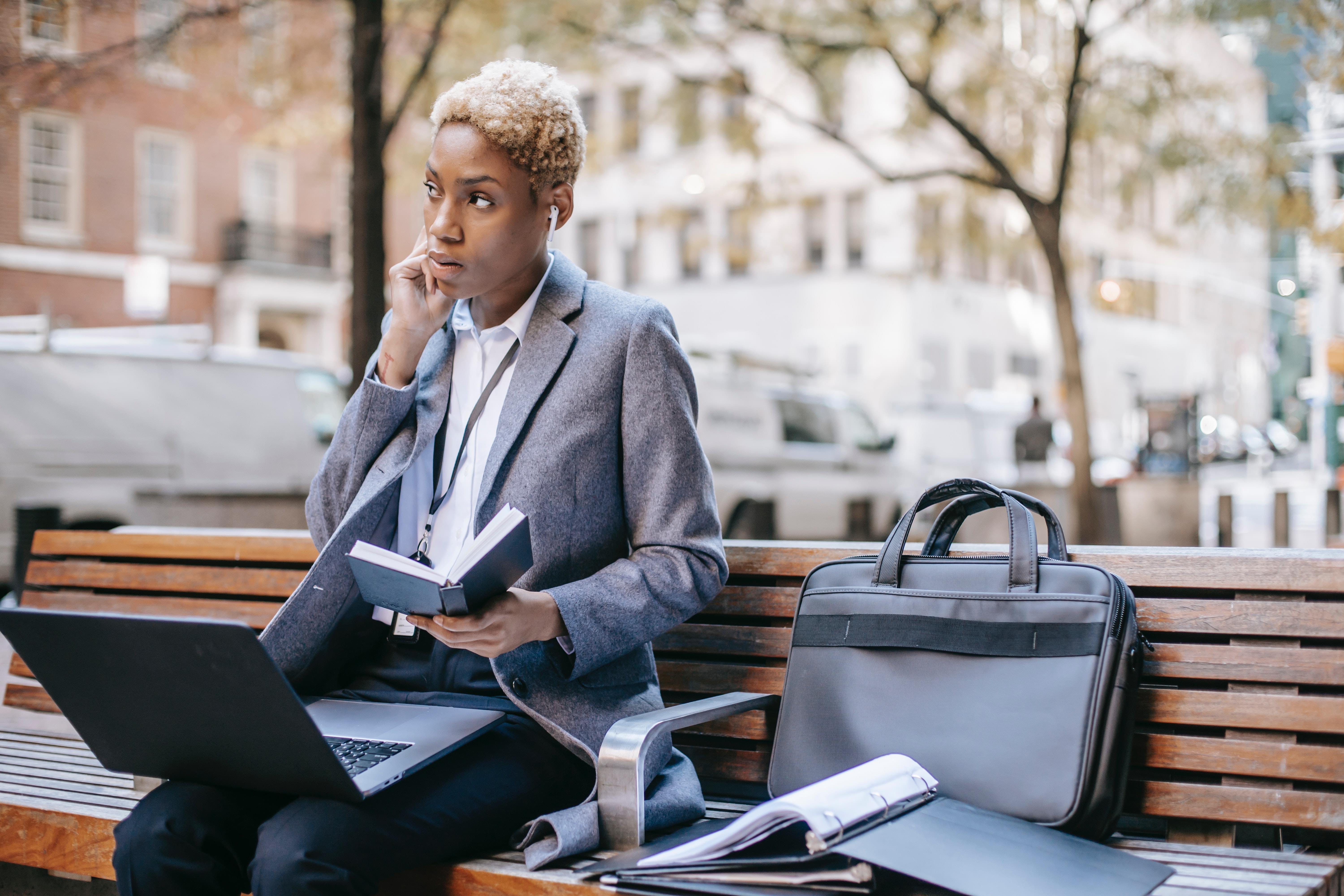 A Black woman seated on a bench with a laptop, cell phone, brief case, and binder