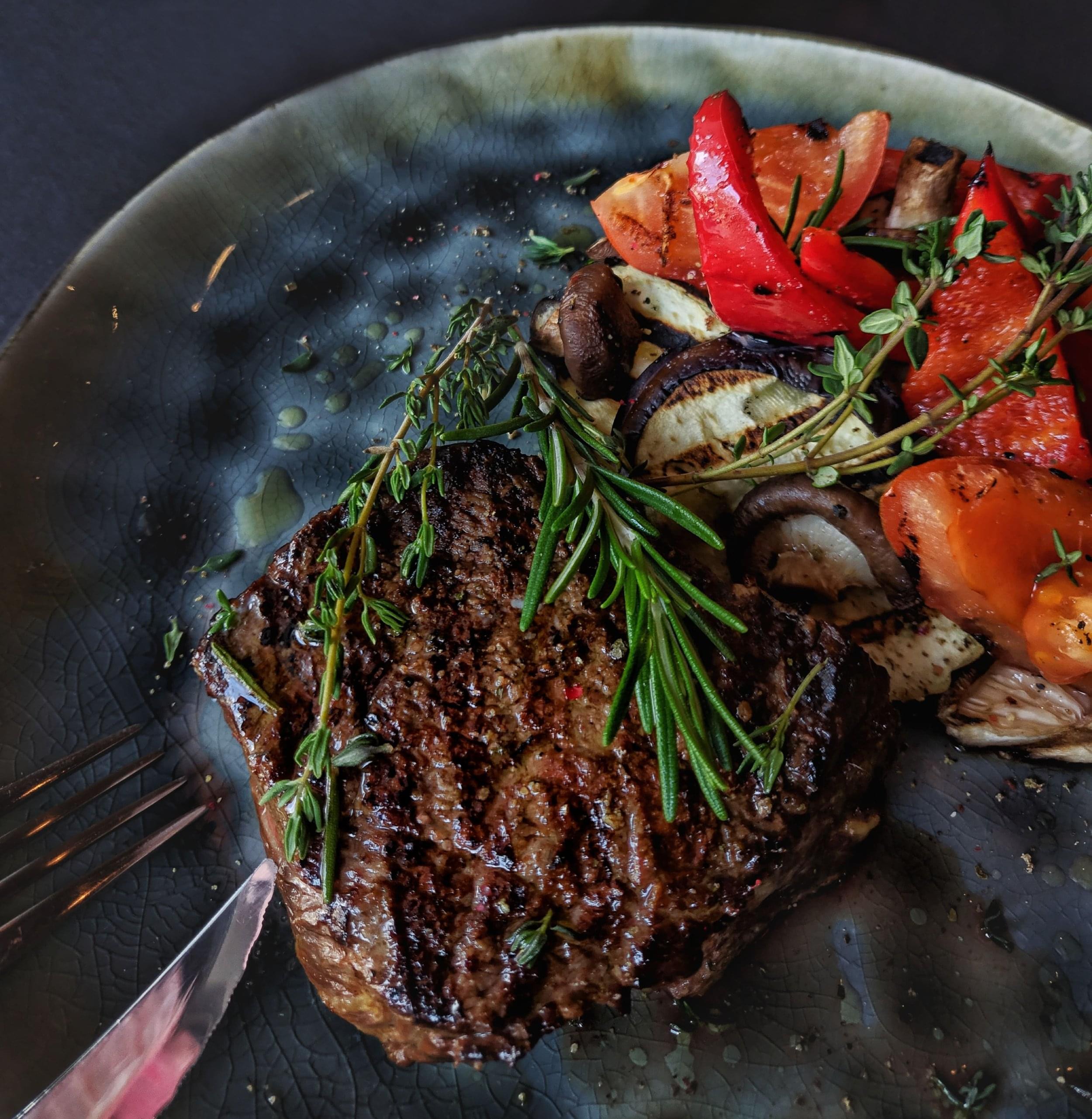 A steak on a plate with sage as garnish, peppers and mushrooms on the side