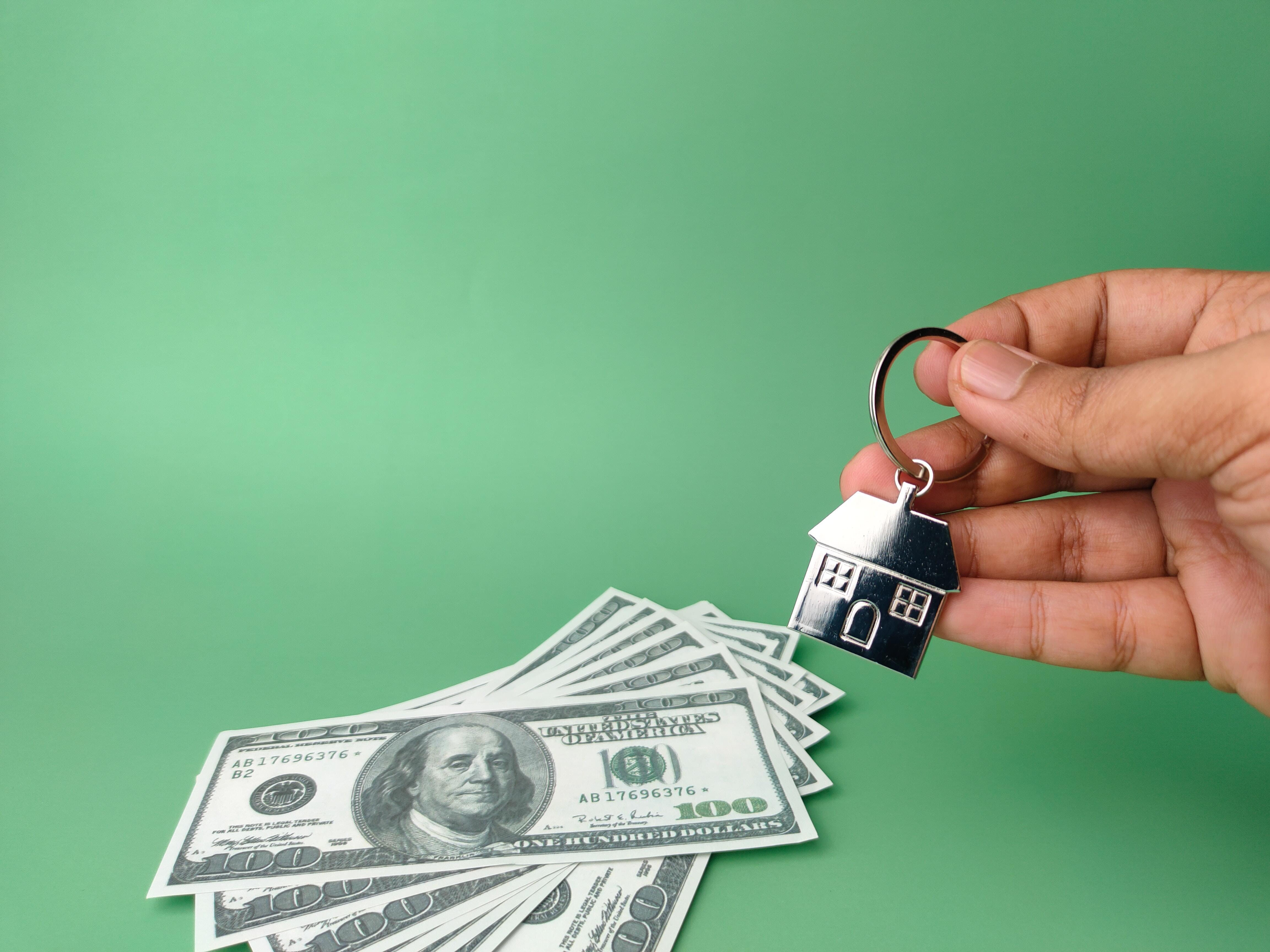 Several hundred-dollar bills on a green background and a hand holding a home-shaped keychain