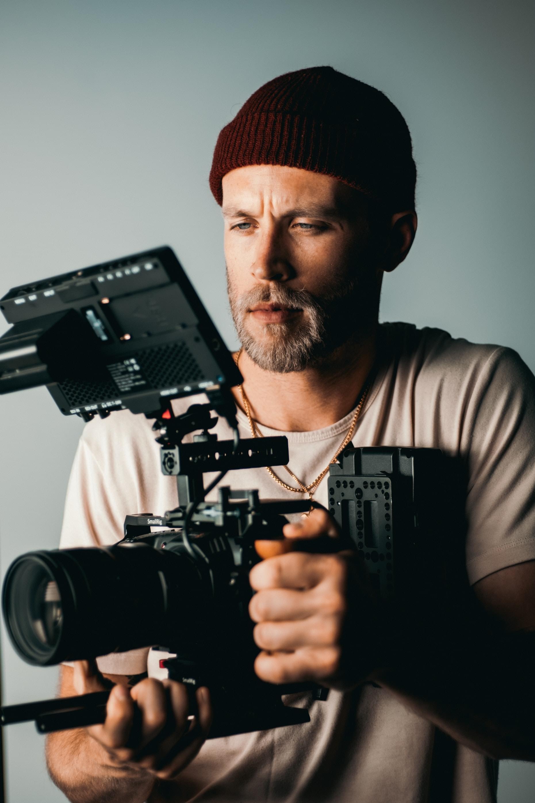 A professional photographer holding a camera, concentrating, wearing a maroon hat in front of a gray background