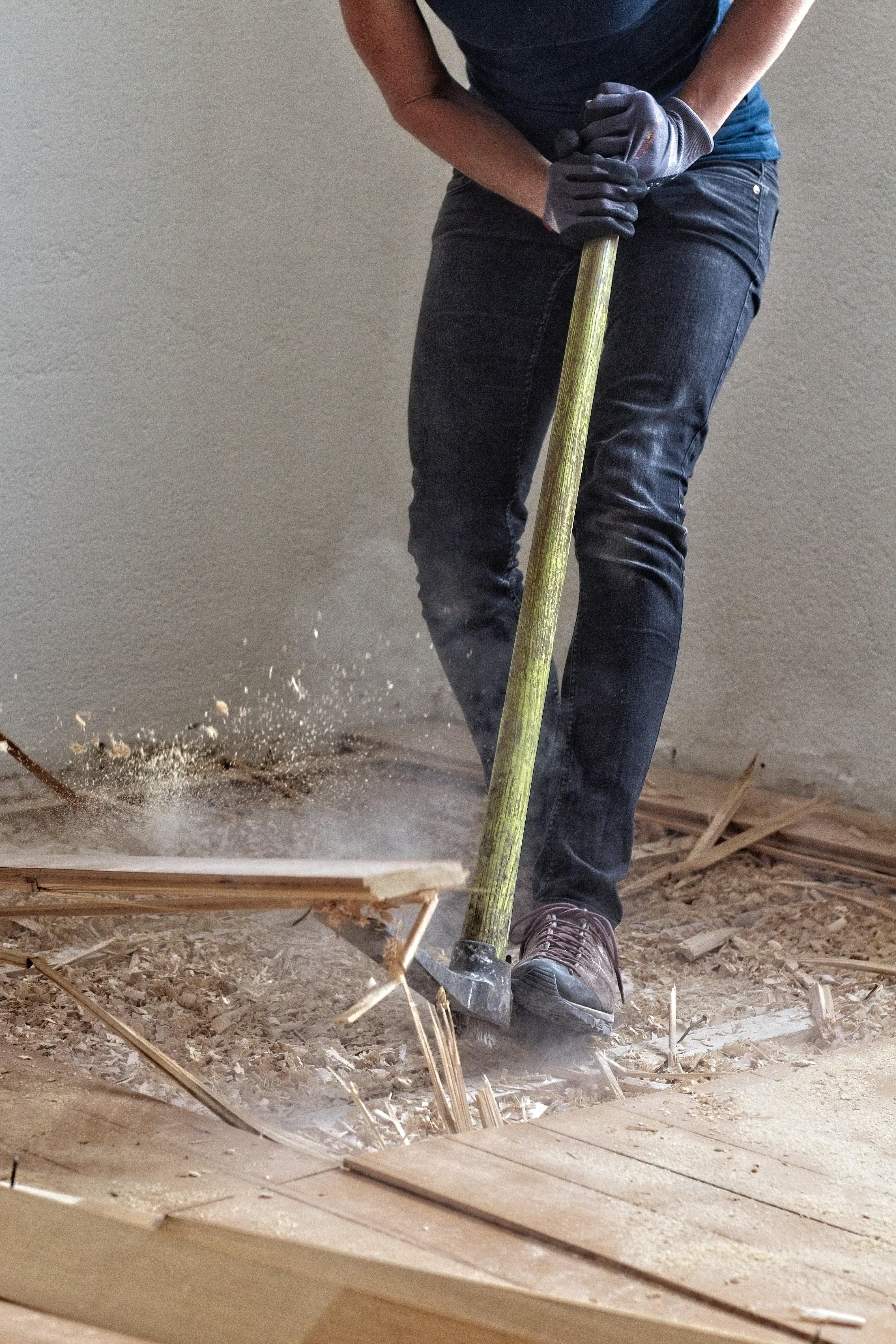 A contractor from the waist down, wearing gloves, prying up old flooring with a pickaxe