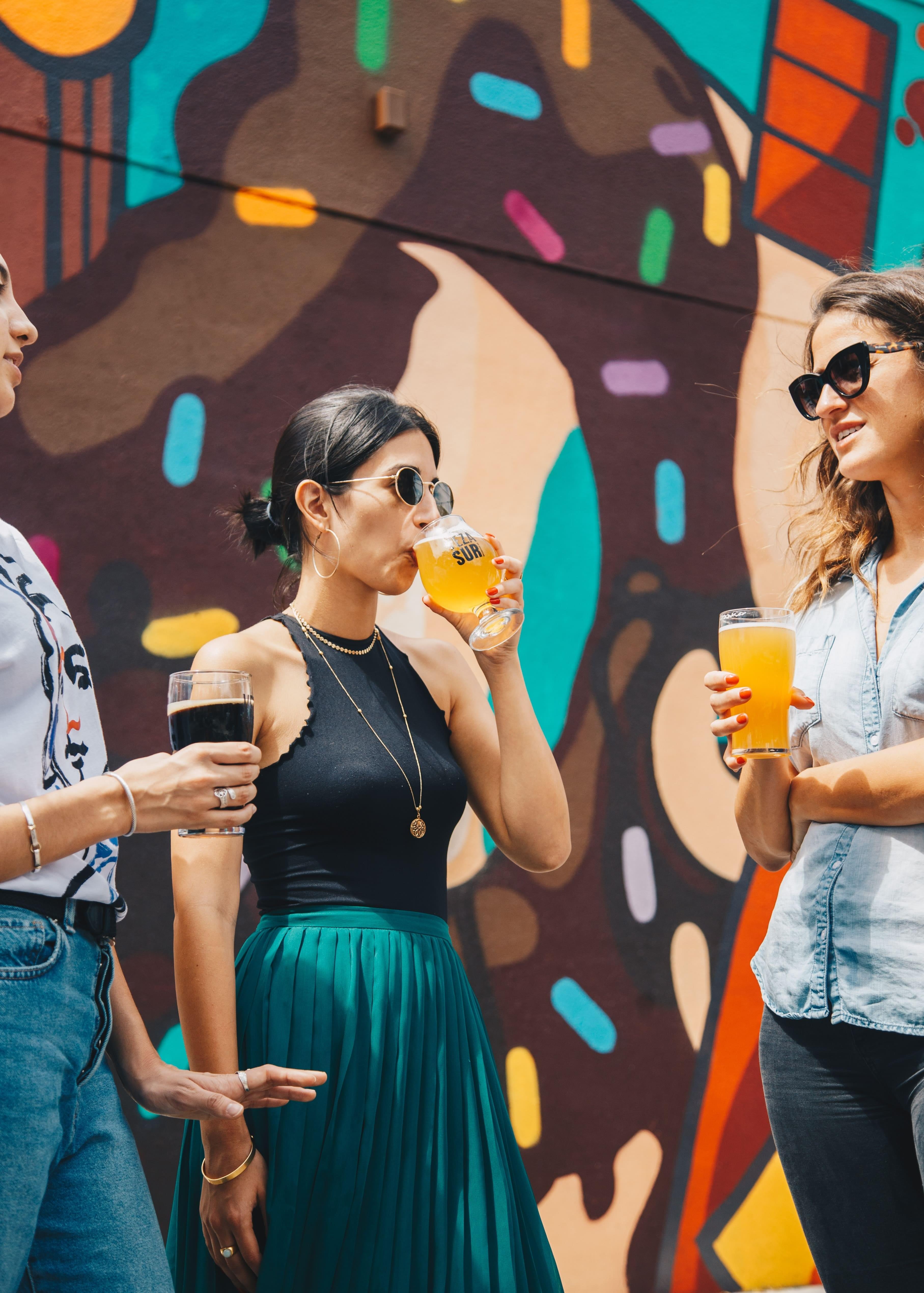 Three dark-haired women standing in front of a huge outdoor mural, socializing and drinking beer