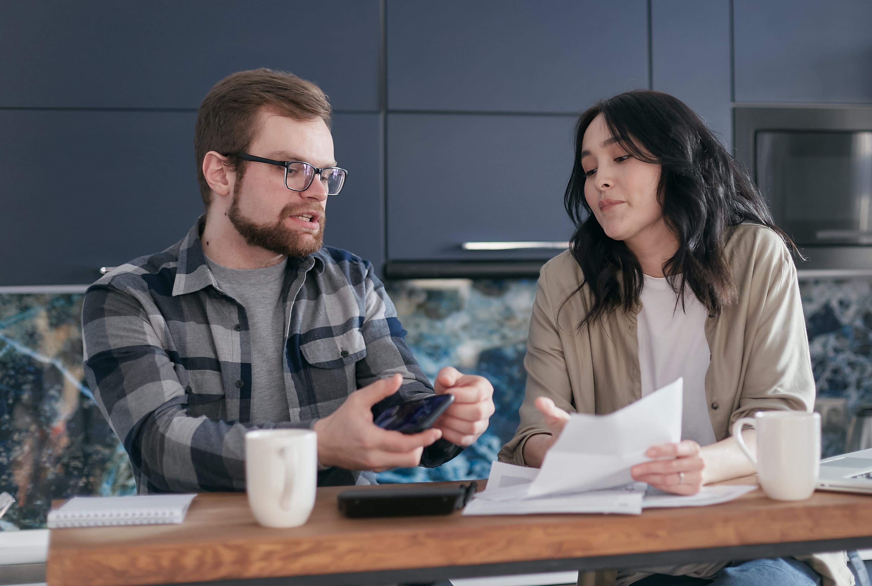A man and woman discussing their financial situation over some paperwork, a phone, and coffee
