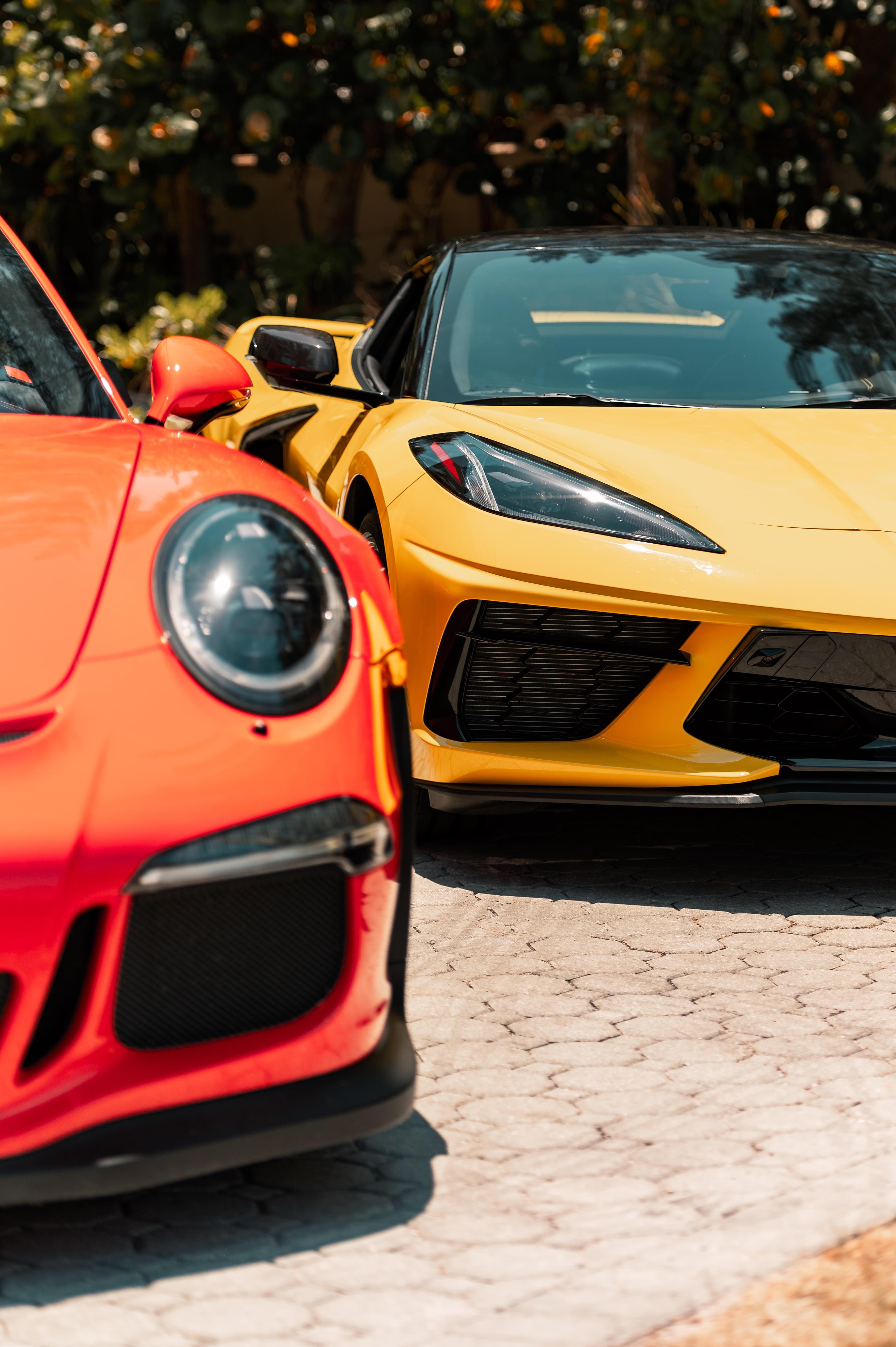 Closeup on one orange and one yellow exotic supercar in front of leafy trees