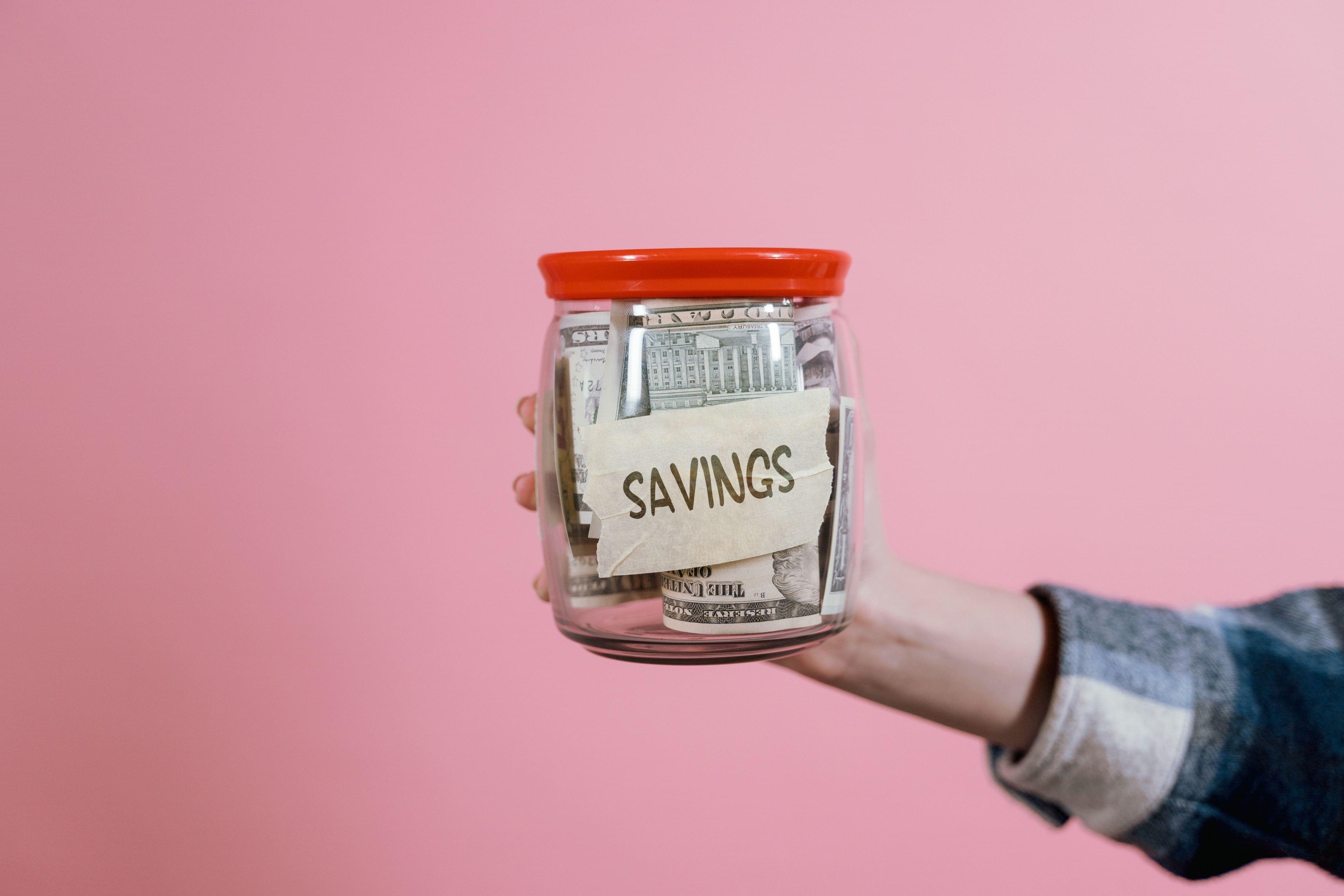 A hand holding a jar full of money with a red cap and a label that says "savings" in front of a pink background