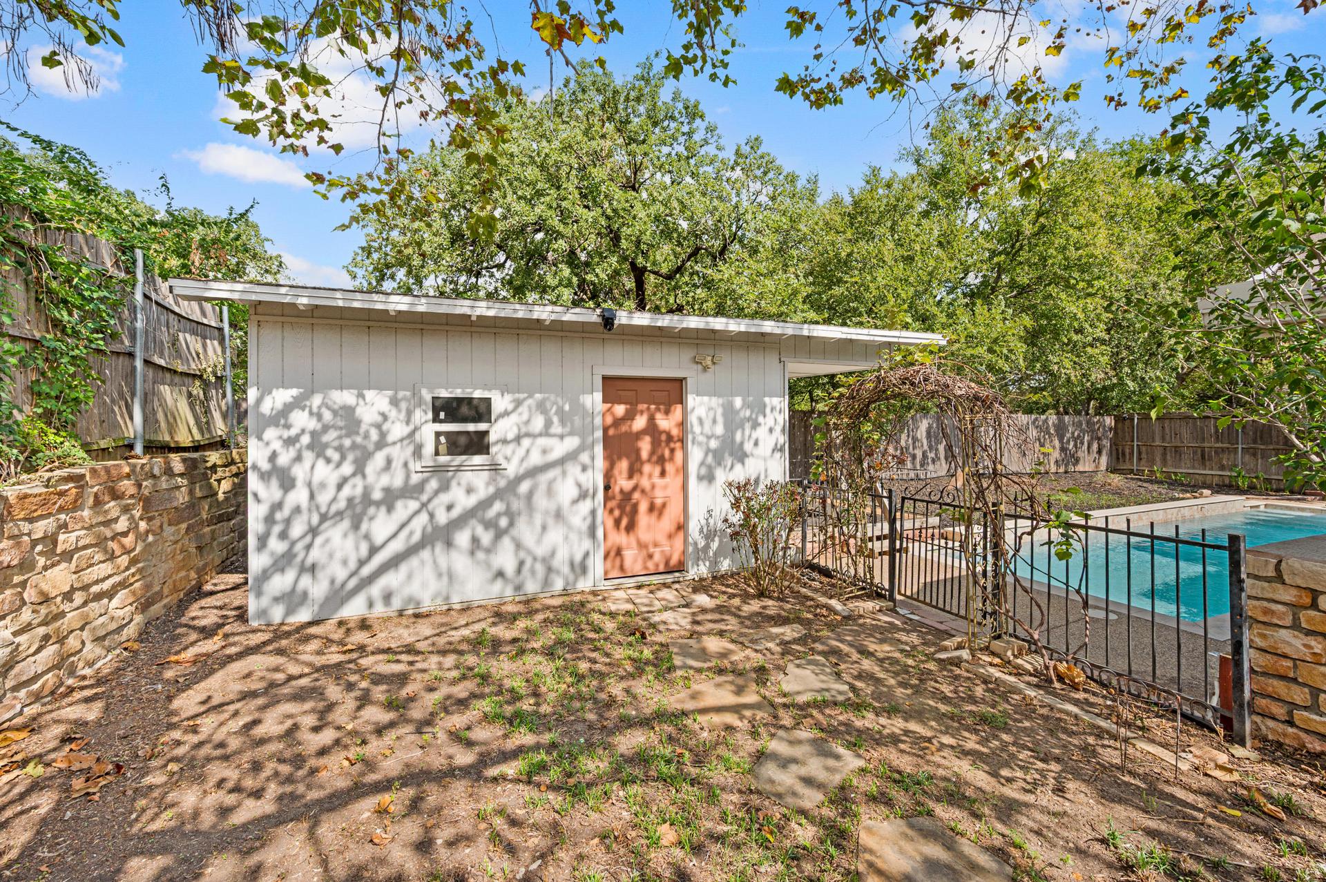 Detached backyard structure with light-colored siding, a small window, and a separate entry door near the pool