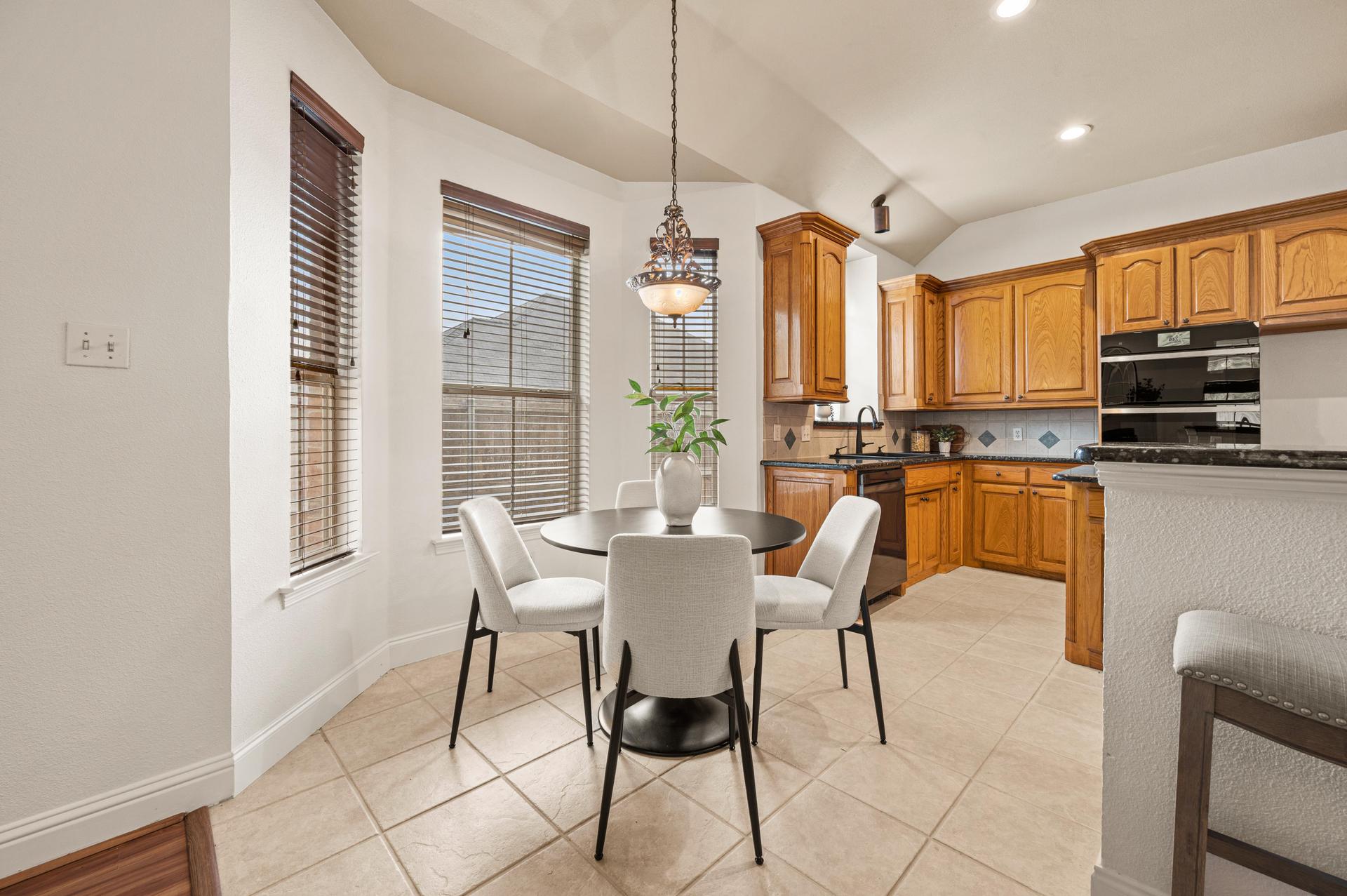 Bright kitchen and dining area with warm wood cabinetry, dark countertops, built-in double ovens, tiled flooring, and large windows.